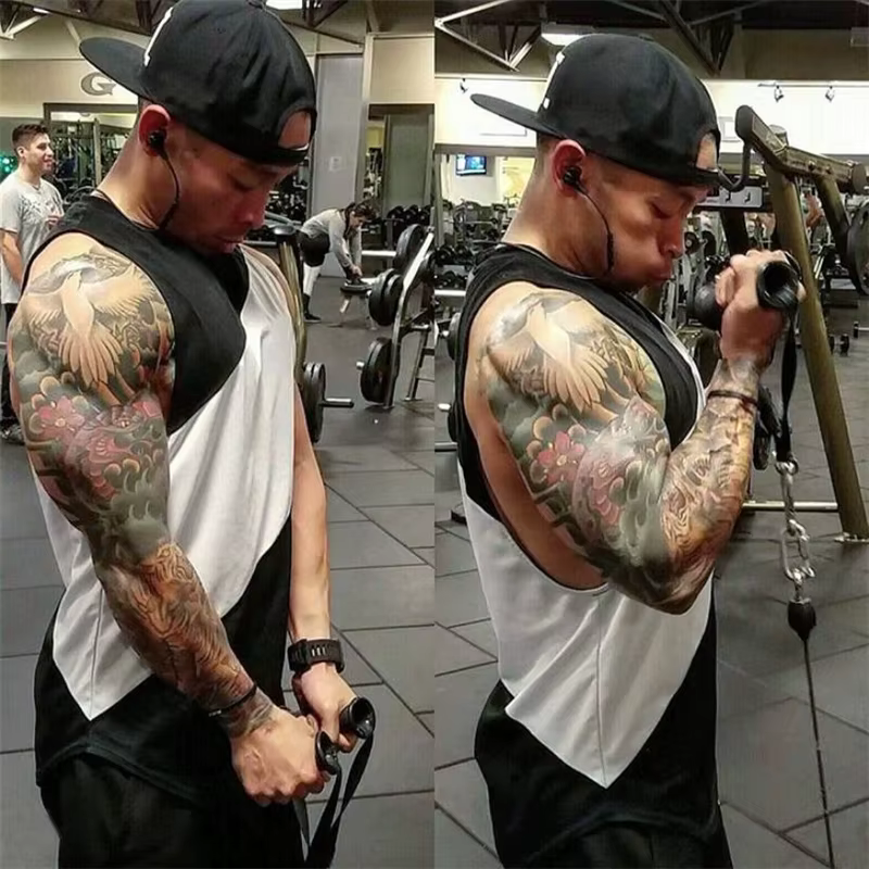 Man in gym wearing a sleeveless tank top, showcasing tattoos while performing a bicep curl on cable machine.