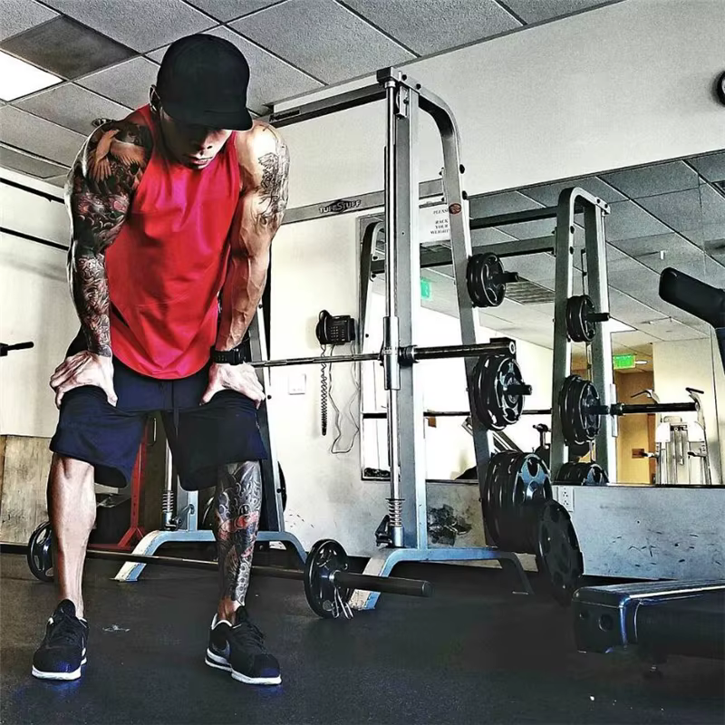 Man wearing a red sleeveless tank top in a gym, focused on workout routine with weights nearby.
