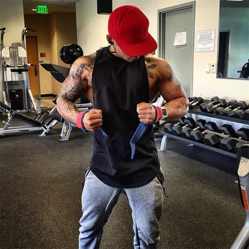 Muscular man wearing a black sleeveless gym tank top and red cap, preparing to lift weights in a fitness center.