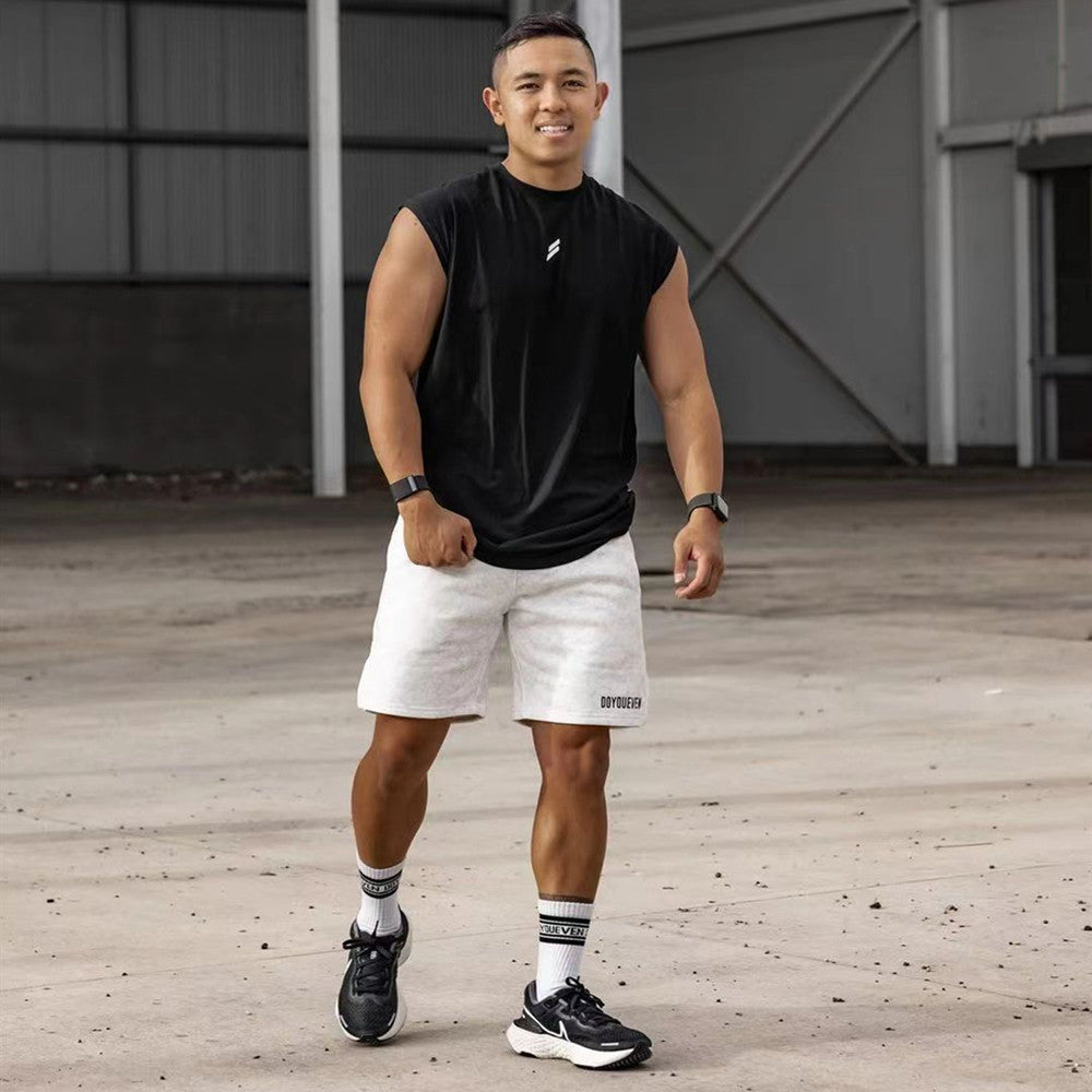 Smiling man in black sleeveless tank top and gray shorts walking in an industrial setting, showcasing athletic wear.