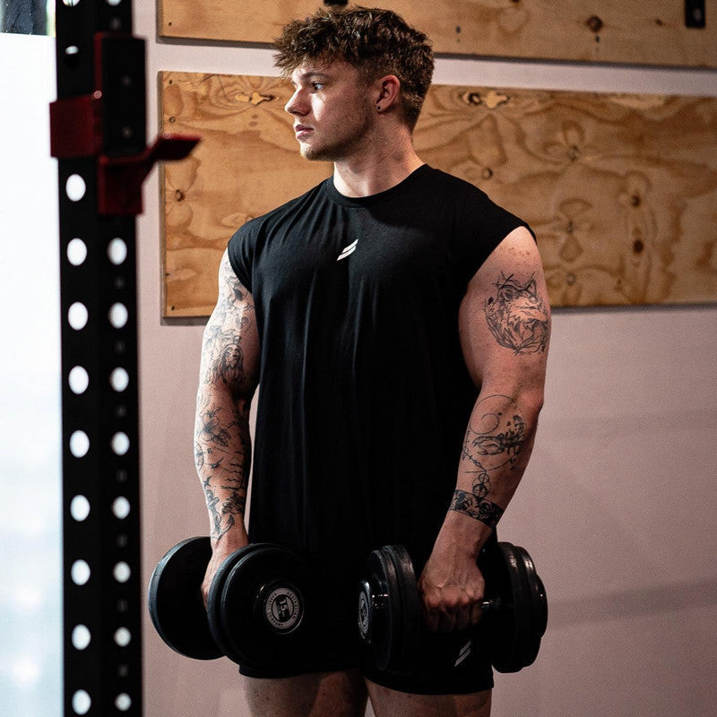 Man lifting heavy dumbbells in a gym, showcasing a muscular build and sporting a sleeveless black tank top.