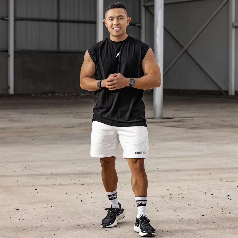 Man wearing a black sleeveless tank top and white shorts, standing confidently in an industrial gym setting.