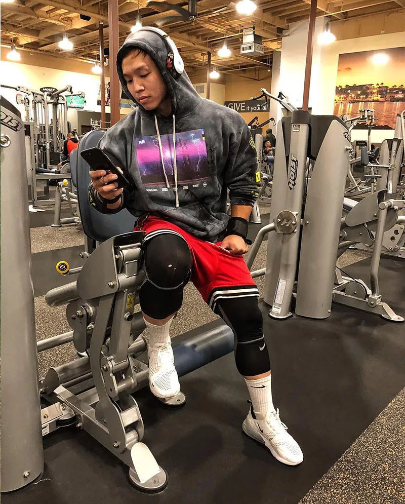 Man in gym wearing a hoodie and basketball shorts, using a fitness machine while checking his phone.