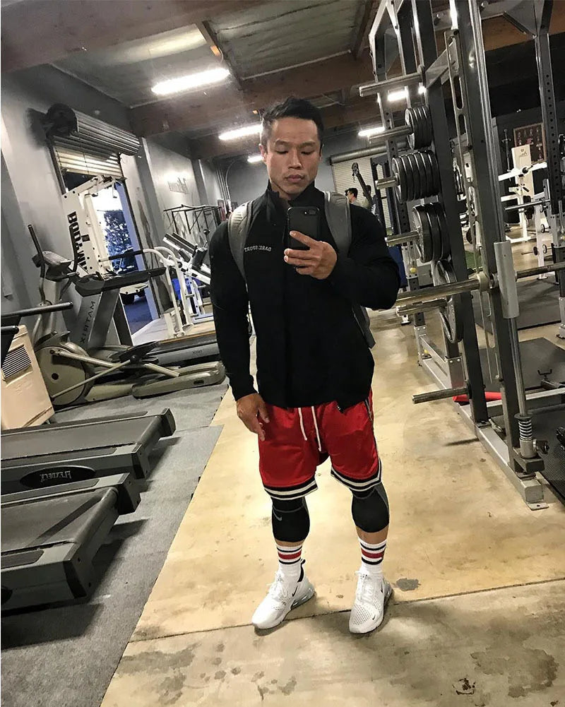 Man in gym wearing black shirt, red basketball shorts, and white shoes, ready for workout in fitness environment.