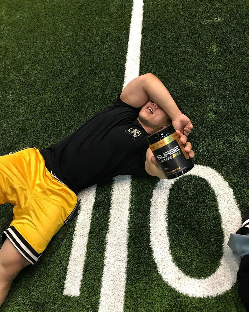 Athlete resting on turf field holding a container of Surge pre-workout in yellow basketball shorts.