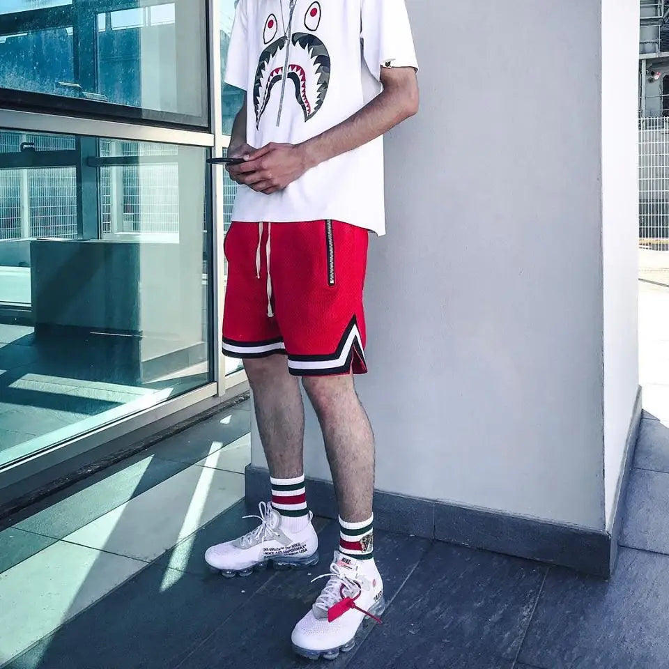 Man wearing red mesh basketball shorts, white shirt, and stylish sneakers, showcasing a casual sporty look.