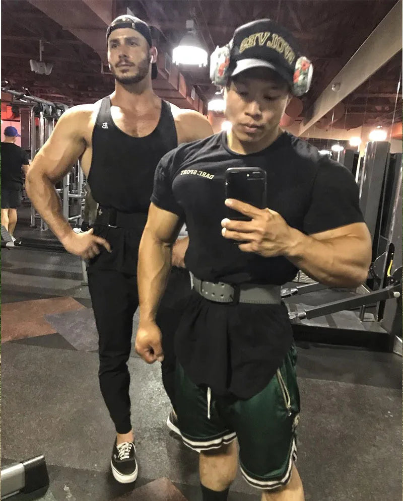 Two men in gym attire posing in a fitness center mirror, showcasing athletic builds and workout gear.