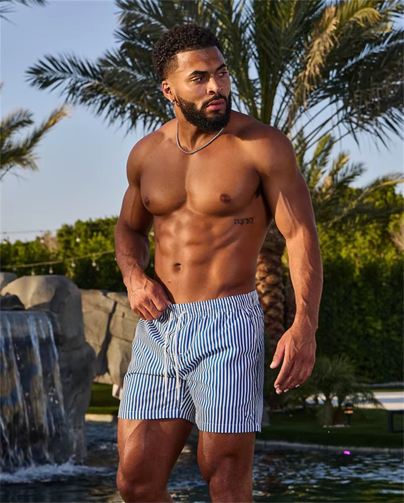 Stylish man wearing striped swim shorts by a pool with palm trees in the background, enjoying a sunny day.