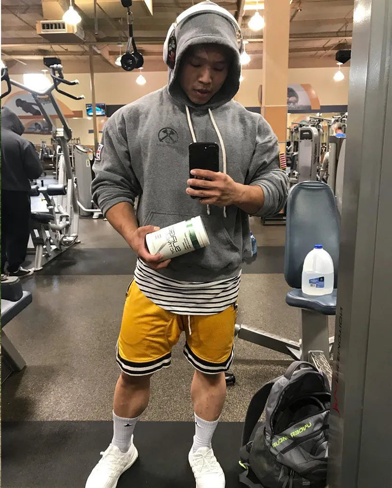 Man wearing gray hoodie and yellow shorts at the gym, holding a sports supplement while looking in a mirror.