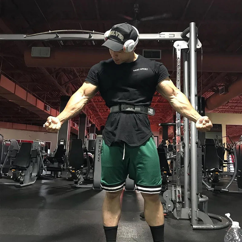 Man showcasing muscular arms in gym, wearing black t-shirt and green mesh basketball shorts, training with intensity.