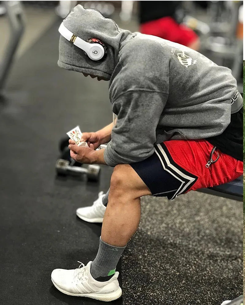 Man in gym wearing headphones, gray hoodie, and red basketball shorts, preparing for workout.